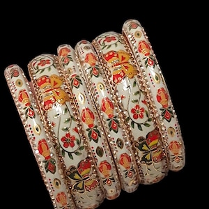 Può includere: Un set di braccialetti bianchi con intricati disegni floreali e a farfalla in rosso, giallo e verde. I braccialetti hanno un bordo color oro e sono su uno sfondo nero. Questi braccialetti decorativi sono un bellissimo accessorio.
