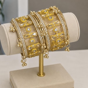 May include: Gold-coloured bangles with clear and yellow detailing, embellished with sparkling rhinestones and dangling charms. The bangles are displayed on a beige jewellery stand, highlighting their intricate design and craftsmanship.