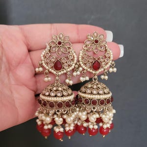 May include: A pair of ornate gold-toned earrings. Each earring features a floral top, a teardrop-shaped red stone, and a bell-shaped bottom. The earrings are adorned with small white pearls and red beads, creating a luxurious look.