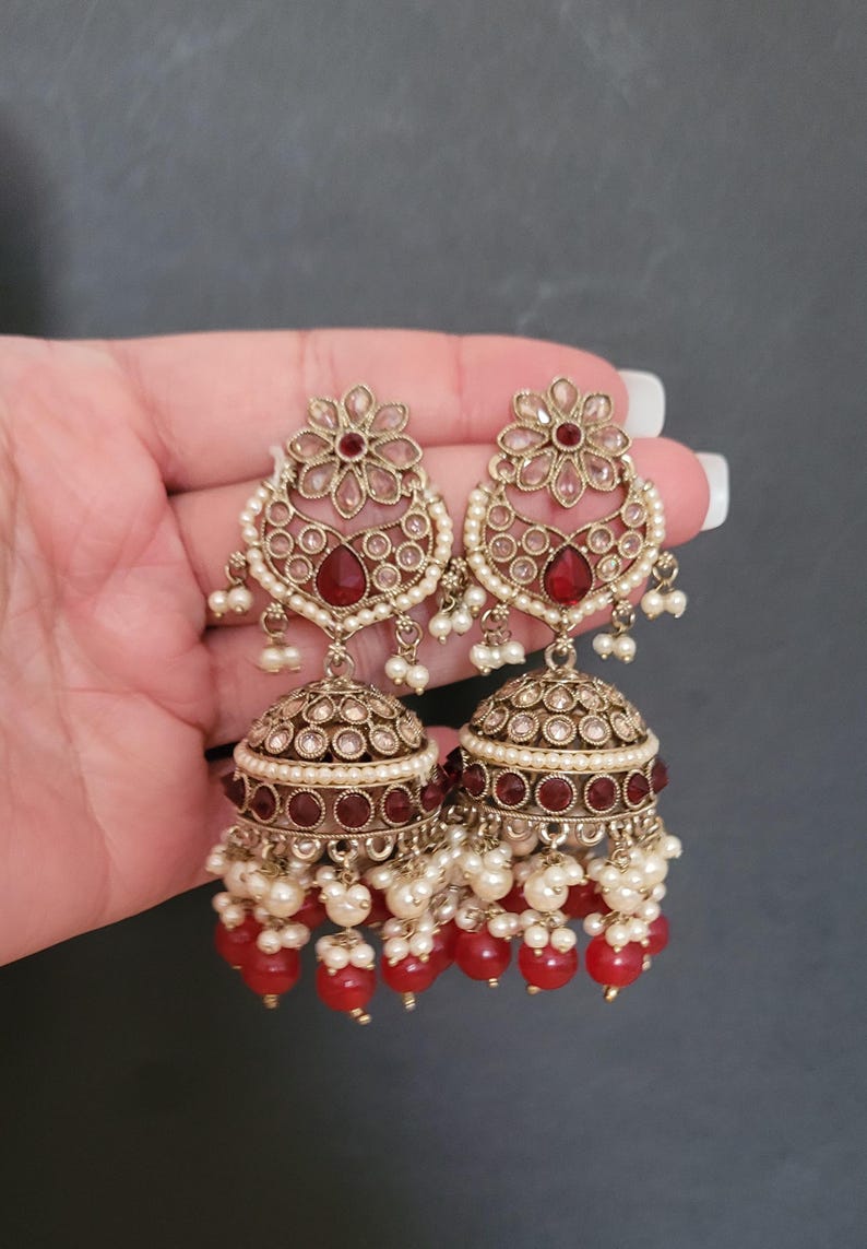 May include: A pair of ornate gold-toned earrings. Each earring features a floral top, a teardrop-shaped red stone, and a bell-shaped bottom. The earrings are adorned with small white pearls and red beads, creating a luxurious look.