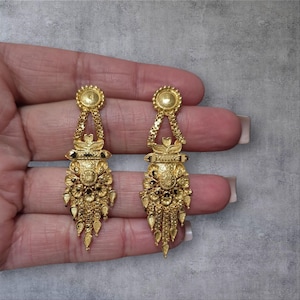 May include: A pair of ornate gold-colored earrings. Each earring features a circular top, a chain-like design, and a detailed, floral-inspired pendant with dangling elements. The earrings are displayed against a gray background.