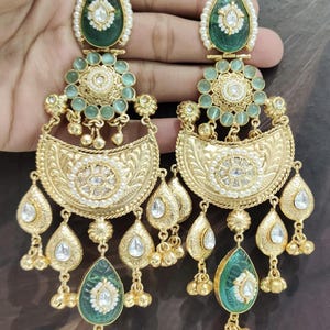 May include: Gold-toned chandelier earrings with intricate detailing. The earrings feature green and white gemstones, pearls, and teardrop-shaped dangles. The design includes floral and crescent-shaped elements, creating an elegant and ornate look.