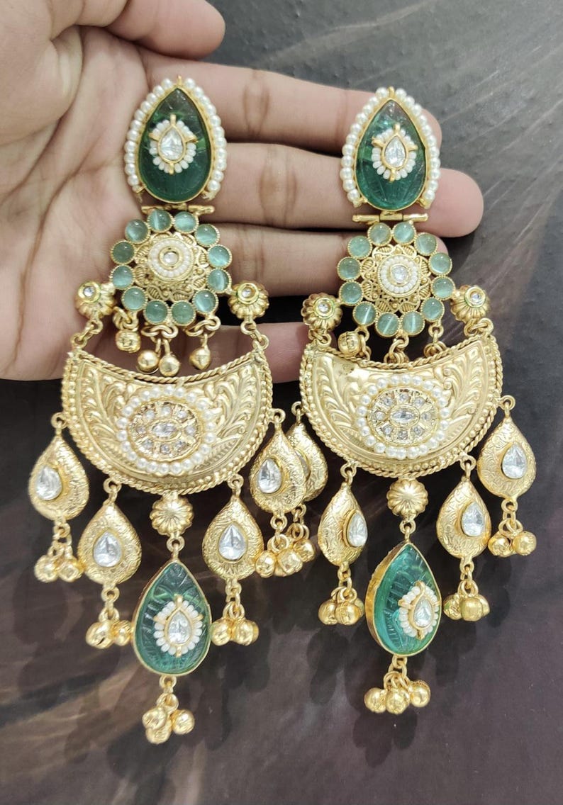 May include: Gold-toned chandelier earrings with intricate detailing. The earrings feature green and white gemstones, pearls, and teardrop-shaped dangles. The design includes floral and crescent-shaped elements, creating an elegant and ornate look.