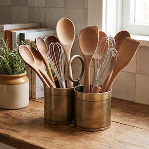 Puede incluir: Una colección de cucharas de madera, espátulas y batidores de alambre metálico guardados en dos recipientes cilíndricos de latón conectados. Los utensilios de cocina están dispuestos verticalmente con cabezales de madera y alambres.