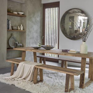 Peut inclure: Une salle à manger rustique avec une longue table en bois et deux bancs assortis. Un plaid crème est posé sur un banc. La pièce comprend un grand miroir rond, un vase blanc avec des branches et un tapis blanc moelleux. La table mesure environ 200 cm de long.