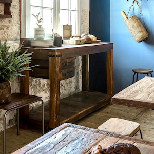 May include: A large, rustic wooden kitchen island with drawers and a lower shelf. The island is stained a warm brown and has metal accents. A woven bag with a baguette hangs on the blue wall. The room has a vintage feel.