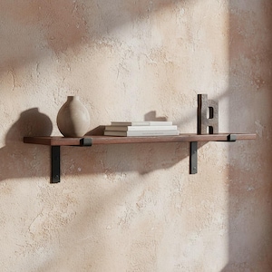 Könnte beinhalten: Ein dunkelbraunes Holzregal mit schwarzen Metallhalterungen, das eine beige Vase, mehrere Bücher und einen dekorativen Buchstaben "H" trägt. Das Regal ist an einer strukturierten, hellbraunen Wand befestigt.