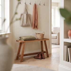 Könnte beinhalten: Eine Holzbank mit einer gefalteten olivgrünen Decke und einem geflochtenen Korb darauf. Eine beige Tragetasche und ein terrakottafarbener Schal hängen an Wandhaken über der Bank, während braune Lederstiefel auf dem Boden darunter stehen.