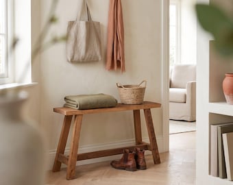 Op de afbeelding: Een houten bank met een gevouwen olijfgroene deken en een gevlochten mand erop. Een beige draagtas en een terracotta-kleurige sjaal hangen aan wandhaken boven de bank, met bruine leren laarzen op de vloer eronder geplaatst.