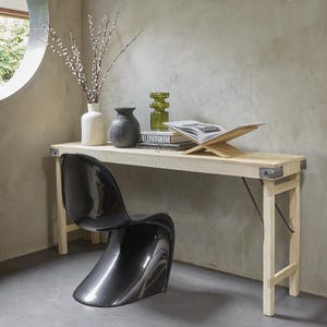 Può includere: Una consolle in legno chiaro con angoli in metallo e una sedia in plastica nera. Il tavolo è decorato con un vaso bianco, un vaso grigio scuro, un vaso di vetro verde e un leggio con un libro aperto. Il tavolo misura circa 120 cm di lunghezza.