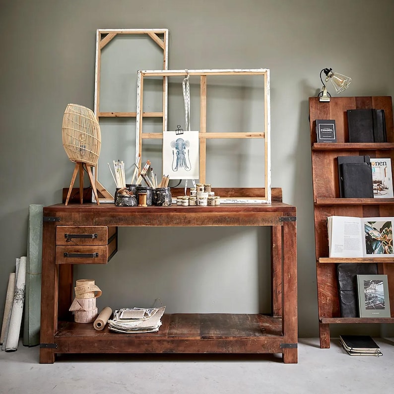 May include: A wooden desk with two drawers, holding art supplies, canvases, and a wicker lamp. A wooden bookshelf is to the right, displaying books and decorative items. The scene is set against a muted green wall.