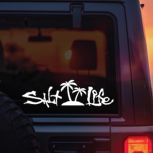 May include: White vinyl decal with the words "Salt Life" in a stylized font, featuring palm trees and an island silhouette. The decal is applied to the rear window of a vehicle, with a sunset visible in the background.