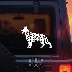 May include: A white decal of a German Shepherd dog silhouette, with the breed name incorporated into the design. The decal is affixed to the rear window of a vehicle. The image is set against a blurred background.