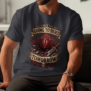 Może przedstawiać: Ciemnoszary t-shirt z nadrukiem graficznym przedstawiającym czerwoną dwudziestościenną kość do gry z widoczną cyfrą jeden. Wzór zawiera sztylet, odłamki kamieni, małą czaszkę oraz napis "THIS IS GOING TO HURT TOMORROW" na wstęgach.