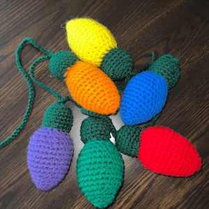 May include: A set of six crocheted lightbulb ornaments in various colors, including yellow, orange, blue, purple, green, and red. The ornaments are connected by a green string.