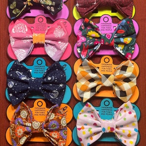 May include: A collection of ten fabric bow ties for pets and people, each mounted on a colorful card labeled Handmade Happiness for Paws and People. The bows feature diverse patterns including paisley, celestial, floral, plaid, and geometric designs.