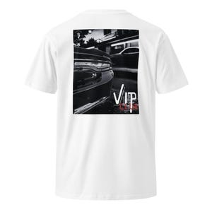 Peut inclure: T-shirt blanc avec un graphisme noir et blanc de l'arrière d'une voiture, avec le mot "VIP" en blanc et rouge. Le t-shirt a un col rond et des manches courtes.