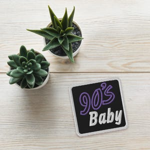 May include: A white square patch with a black background and purple text that reads "90's Baby".