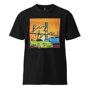 Puede incluir: Camiseta negra con un diseño gráfico del Golden Gate Bridge y la frase "Bay Area". Dos coches, uno verde y otro azul, frente al puente sobre un atardecer naranja. Diseño retro.