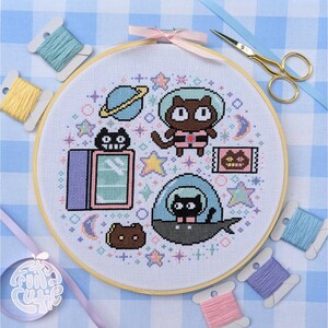 May include: A round wooden embroidery hoop containing white fabric with a cross-stitch pattern of two cats in space suits, a planet, stars, moons, and a cat-shaped television. Gold-tone scissors and five spools of embroidery floss are nearby.