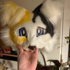 Könnte beinhalten: Eine pelzige Katzenmaske mit weißem Gesicht, gelben und schwarzen Akzenten und blauen Augen. Die Maske hat flauschiges weißes Fell um die Ohren und Wangen sowie eine braune Nase und Zähne. Die Maske wird von einer Hand gehalten.