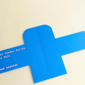 Puede incluir: Una tarjeta azul brillante, troquelada con forma de cruz, con texto blanco. La tarjeta está sobre un fondo crema. El texto incluye las palabras "ni Cüzdan Kalıbı", "x Tela" y "hnk atelier".