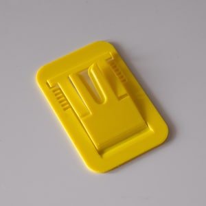 May include: A bright yellow plastic clip, rectangular in shape, with a raised central section and a U-shaped opening. The clip has small, ridged details along the sides, and is designed to hold items together. The clip is shown against a white background.