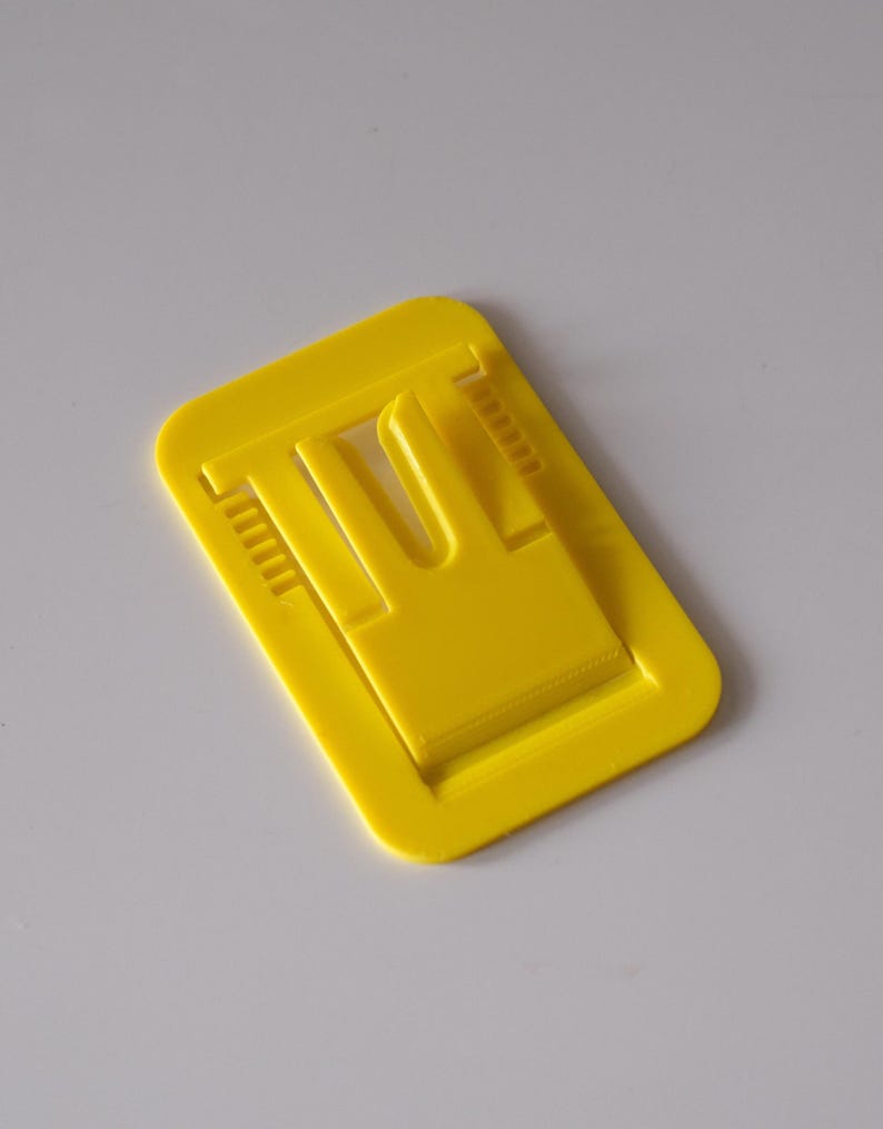 May include: A bright yellow plastic clip, rectangular in shape, with a raised central section and a U-shaped opening. The clip has small, ridged details along the sides, and is designed to hold items together. The clip is shown against a white background.