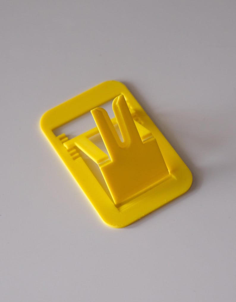 May include: A bright yellow plastic phone stand with a rectangular base and a built-in support. The stand has a slot to hold a phone upright. The design is simple and functional, suitable for desks or tables.