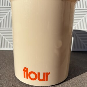 May include: A beige plastic container with a wide mouth and a rounded bottom. The container has the word "flour" printed in orange on the front.