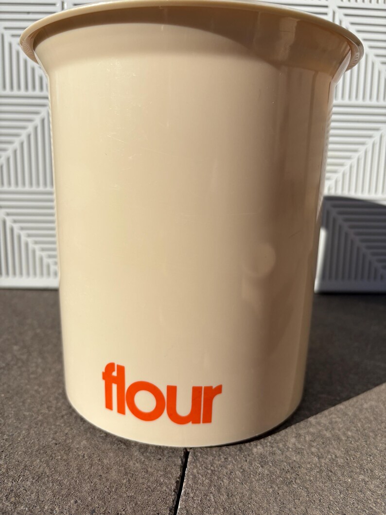 May include: A beige plastic container with a wide mouth and a rounded bottom. The container has the word "flour" printed in orange on the front.