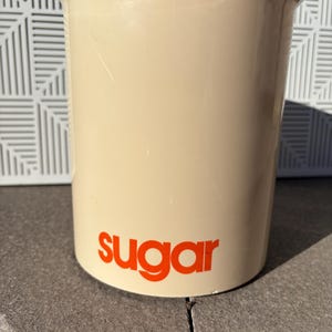 May include: A beige ceramic sugar container with the word "sugar" printed in orange on the front.