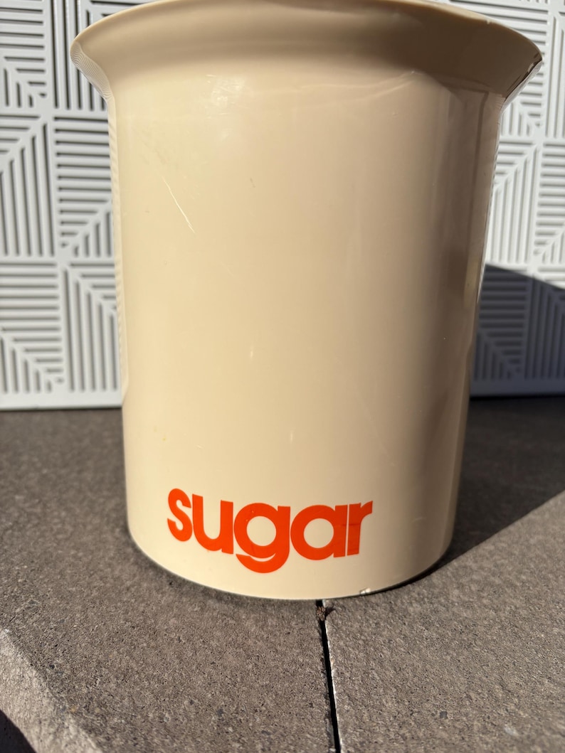 May include: A beige ceramic sugar container with the word "sugar" printed in orange on the front.