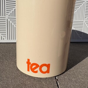May include: A white ceramic tea canister with the word "tea" printed in orange.