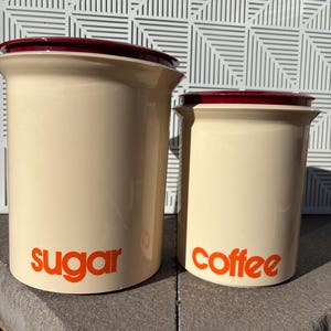 May include: Two ceramic canisters with red lids. The larger canister is labeled "sugar" and the smaller canister is labeled "coffee".