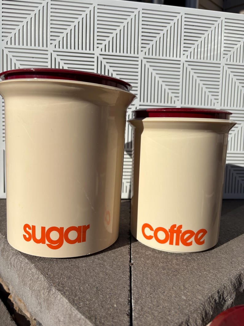 May include: Two ceramic canisters with red lids. The larger canister is labeled "sugar" and the smaller canister is labeled "coffee".