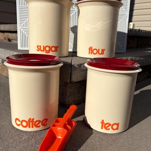 May include: Set of four vintage ceramic canisters with red lids and orange lettering. The canisters are labeled "sugar", "flour", "coffee", and "tea".  A set of orange plastic scoops is included.