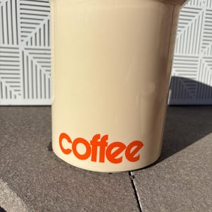 May include: A beige ceramic coffee canister with the word "coffee" printed in orange on the front.