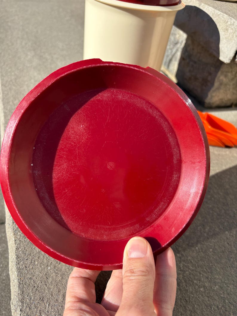 May include: A red plastic lid for a container. The lid is round and has a smooth surface.