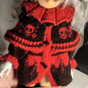 May include: A doll with long silver hair wearing a knitted red and dark brown jacket. The jacket features a ruffled collar, a skull pattern on the shoulders, and a button closure. The doll has large eyes and a pale complexion with pink blush.