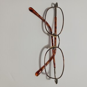 May include: Eyeglasses with oval lenses and a thin, silver-colored metal frame. The temples are a tortoiseshell pattern in shades of brown and amber. The glasses are set against a plain white background.
