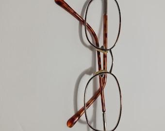 May include: Eyeglasses with oval lenses and a thin, silver-colored metal frame. The temples are a tortoiseshell pattern in shades of brown and amber. The glasses are set against a plain white background.
