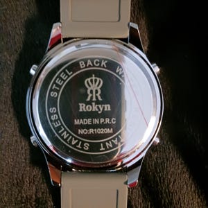 May include: A stainless steel watch with a gray band. The watch face is silver with the text "STAINLESS STEEL BACK WATER RESISTANT" around the edge. The center of the watch face has the brand name "Rokyn" and "MADE IN P.R.C" and "NO:R1020M".