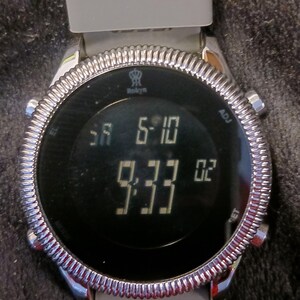 May include: A digital wristwatch with a gray band and a silver-colored bezel. The watch face displays the time as 9:33 and 6:10. The brand name "Rokyn" is visible on the watch face.