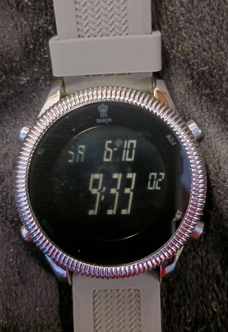 May include: A digital wristwatch with a gray band and a silver-colored bezel. The watch face displays the time as 9:33 and 6:10. The brand name "Rokyn" is visible on the watch face.