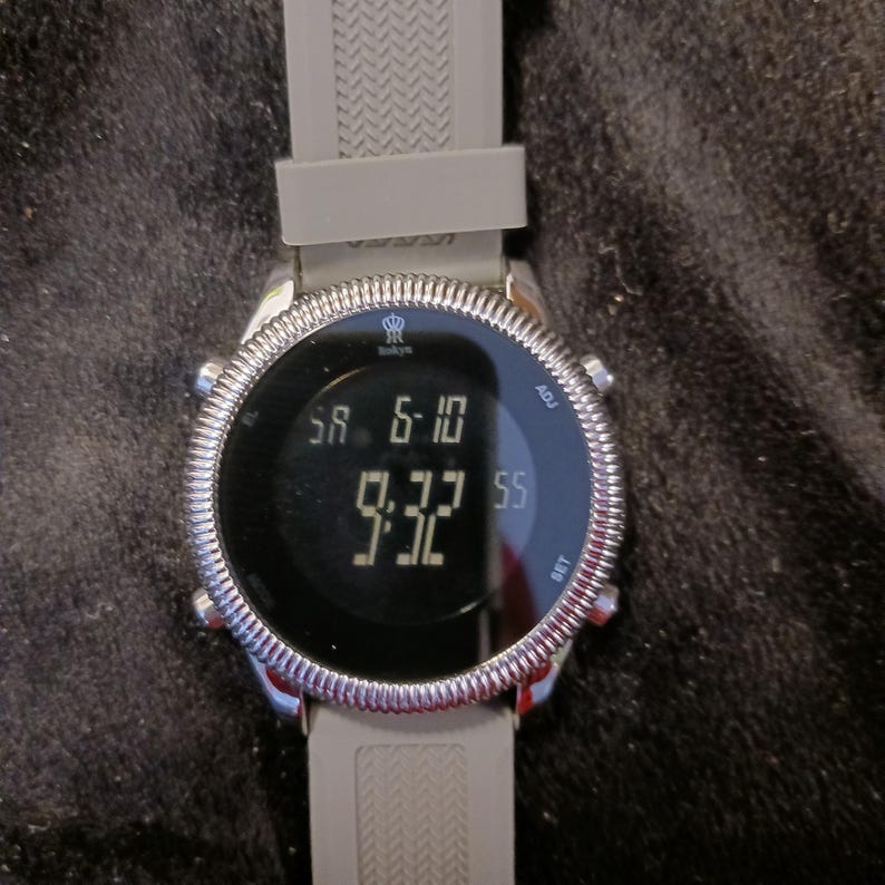 May include: A digital wristwatch with a gray silicone band and a silver-colored bezel. The watch face displays the date as SA 6-10 and the time as 9:32. The watch has buttons labeled ADJ and SET.