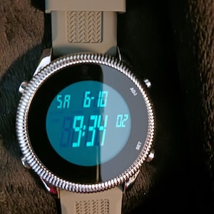 May include: A digital wristwatch with a silver bezel and a gray band. The watch face displays the date and time in blue digits. The time reads 9:34:02. The watch has buttons labeled "ADJ" and "SET".