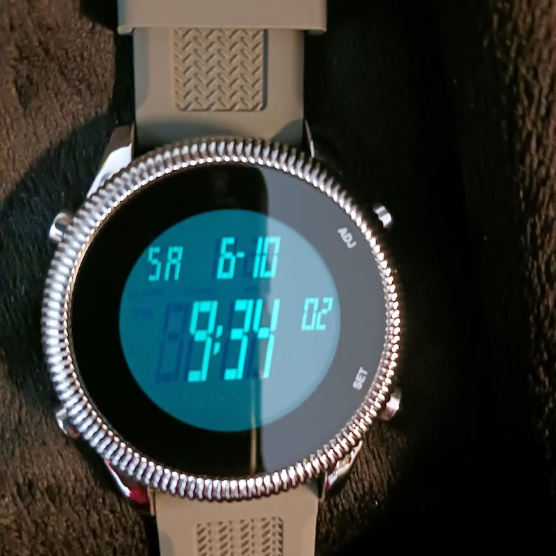 May include: A digital wristwatch with a silver bezel and a gray band. The watch face displays the date and time in blue digits. The time reads 9:34:02. The watch has buttons labeled "ADJ" and "SET".