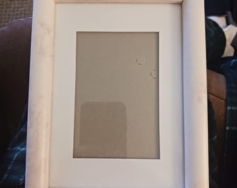 Puede incluir: Un marco de fotos rectangular blanco con un borde rosa-marrón claro. El marco tiene un paspartú blanco y una cubierta de vidrio o plástico transparente. Diseñado para una fotografía o una obra de arte.