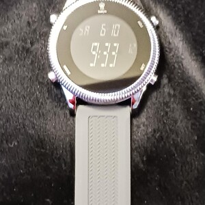 May include: A digital wristwatch with a gray silicone band. The watch face displays the date as 'SA 6 10' and the time as '9:33'. The watch has a silver-colored bezel and a blue clasp.